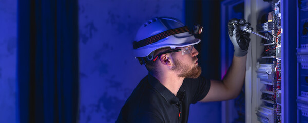 A male electrician works in a switchboard in overalls against the backdrop of emergency lighting.
