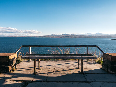 Empty old wooden bench seat at Sevanavank Monastery with Lake Sevan view with mountains landscape and blue sky background, Armenia country. Public seating for taking a break and enjoying the view.