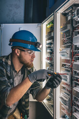 A male electrician works in a switchboard in overalls against the backdrop of emergency lighting.