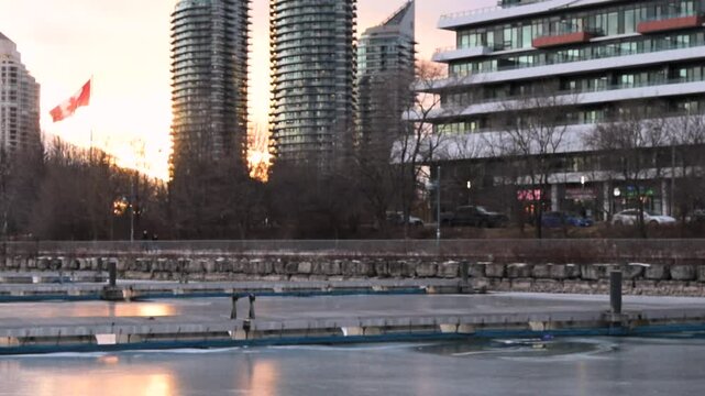 A tranquil sunset at Toronto&rsquo;s waterfront near Mimico and Lake Shore. High-rise buildings glow in the golden light as ducks swim peacefully in the calm water, creating a serene urban scene.
