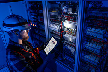 Electrician in overalls, focused on work in switchboard with fuses, using tablet. Top view.