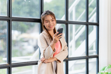 Smiling Woman using Smartphone by Window: A young woman smiles gently as she uses her smartphone, standing casually by a large window in a contemporary setting.
