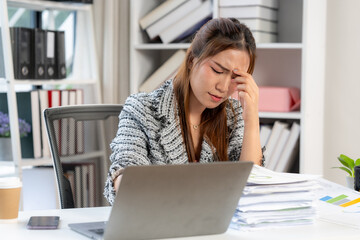 Overwhelmed Businesswoman: A young businesswoman sits at her desk, hand to her forehead, looking stressed and overwhelmed by paperwork and a laptop.  The image depicts the pressures of modern work. 