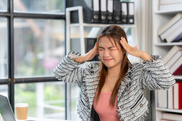 Overwhelmed Businesswoman: A young businesswoman sits at her desk, hands pressed to her temples,...