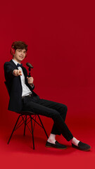 Obraz premium Smiling teen boy, student in black suit and red headphones, sitting on chair with microphone, recording online podcast against red studio background. Concept of youth, education, emotions