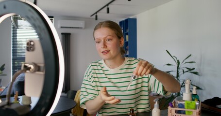 A young woman shoots a beauty blog at home. She smiles and talks while applying cosmetic lotion to...