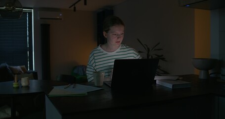 A young woman uses a laptop while sitting at a table in the dark. Notebooks and books on the table. Preparing for the session at night