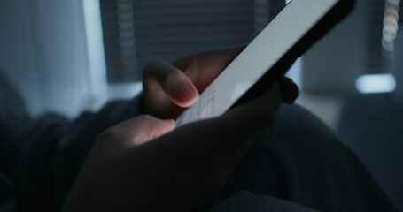 A woman uses a mobile phone while sitting in a dark room. Close-up of her hands, an unrecognizable...