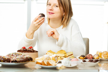 The pretty woman eats a sweet dessert in front of table with cakes and tarts. Concept of sugar addict people who can't resist eating sweets, diabetes