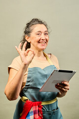 Fototapeta premium Smiling senior woman wearing apron showing ok sign and holding tablet