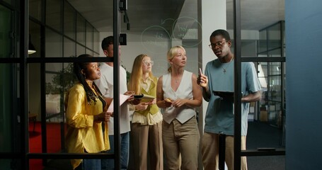 A group of young people of different nationalities are talking standing near a glass board with notes. Business planning. Daylight