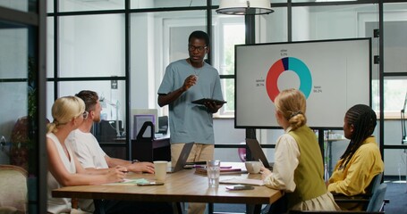 A speaker with a tablet in his hand talks standing in front of his colleagues at an office meeting. Analytical graphs on the screen. Business planning.