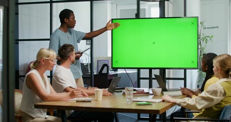 A man with a tablet in his hand is talking standing near a green screen at an office meeting. His colleagues are sitting at the table. Copy space, mockup