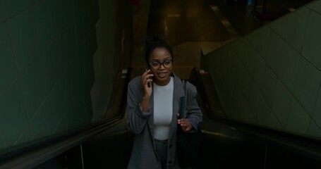 A woman in formal clothes is talking on a mobile phone. She rides the escalator coming up from the subway station