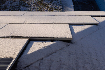 Snow-covered solar panels on a rooftop illuminated by sunlight, with a frosty backyard visible in...