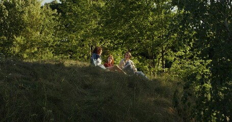Three friends are smiling and talking sitting on the grass on the edge of the forest. A sunny summer day