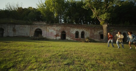 Obraz premium Three young friends are having fun and running around the ruins of a historic building. The fortress walls are overgrown with green vegetation. A sunny summer day