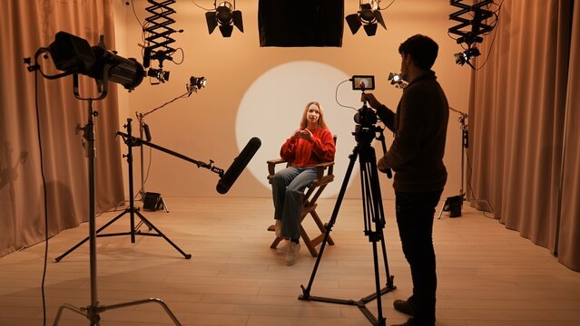 Studio for photo video production filming with lighting equipment and cyclorama, model actress sits in front of camera on chair and talking, recording interview.
