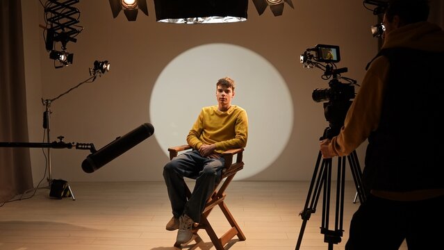 Studio for photo video production filming with lighting equipment and cyclorama, model actor sits in front of camera on chair and talking, recording interview.