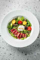 A beautifully arranged salad featuring seared tuna, fresh greens, cherry tomatoes, and roasted potatoes, topped with a poached egg and a sprinkle of nuts, served in a stylish white bowl.
