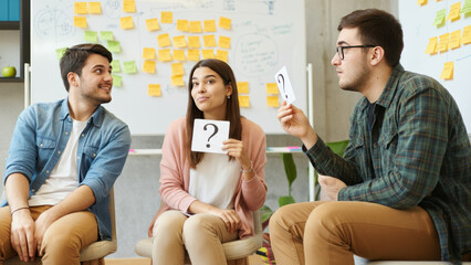  Group of young coworkers participating in a team-building quiz with question cards, collaborating in a modern office with sticky notes on the wall.