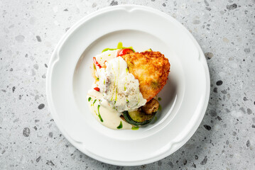 A beautifully presented dish featuring a crispy, breaded chicken fillet topped with creamy sauce, served alongside sautéed vegetables and garnished with a drizzle of fresh herbs.