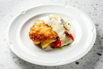 A beautifully presented dish featuring a crispy, breaded chicken fillet topped with creamy sauce, served alongside sautéed vegetables and garnished with a drizzle of fresh herbs.