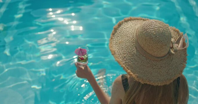 Woman in a straw hat relaxing by a swimming pool, holding a cocktail with a pink umbrella, enjoying a sunny vacation.