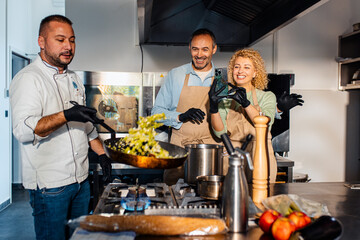 Couple enjoying a cooking class with a chef preparing meal in the kitchen, woman recording with smart phone.