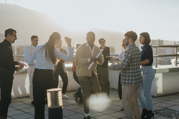 A group of multiracial business colleagues is seen enjoying a celebration at sunset. They are dancing and socializing on a high-rise balcony, symbolizing success and teamwork in a relaxed corporate