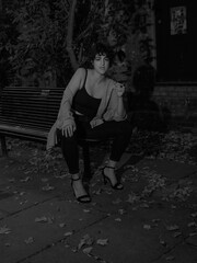 A young woman in a white shirt and dark pants stands against a brick wall at night, under soft monochrome lighting, surrounded by fallen leaves