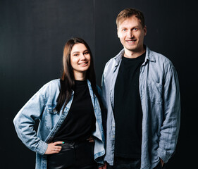 Couple smiles in dark backdrop. Young man and woman stand together, both wearing casual outfits, smiling brightly while posing for a portrait.