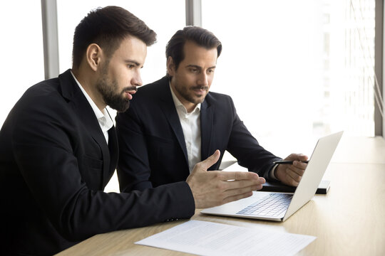 Two businessmen wear suits sit at table with laptop, discussing details of project, reviewing progress, brainstorming ideas, preparing for upcoming presentation, looking at financial data or reports