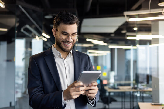 Successful businessman in business suit holding tablet computer. Man inside office at workplace smiling happily, using financial investment app.