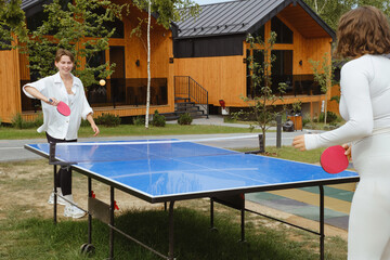 Two women play an outdoor game of table tennis, surrounded by wooden cabins and trees. Summer Sunny...