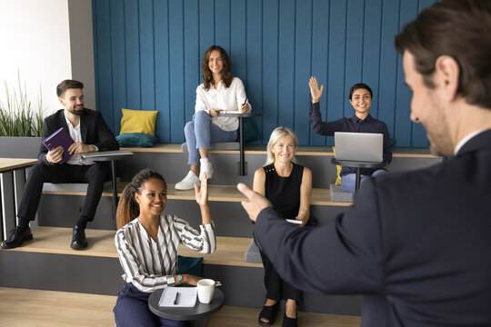Young African woman raise her arm voting during seminar, educational training or briefing event seated with teammates in auditorium, smile, look at male trainer, coach standing in front of audience
