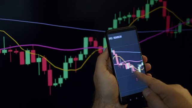 Trader or investor monitoring or looking stock market or crypto market graphics or charts on phone and PC. Man&rsquo;s hand holding phone with graphics in front of PS screen.