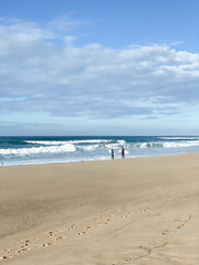 Sandy beach with ocean waves, two surfers walking toward the water with surfboards,