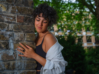 A young woman with curly hair and a white blouse peeks from behind a brick wall in an urban garden, her gaze contemplative and serene in the soft evening light