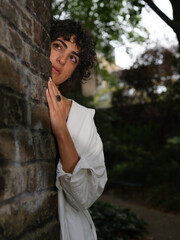 A young woman with curly hair and a white blouse peeks from behind a brick wall in an urban garden, her gaze contemplative and serene in the soft evening light