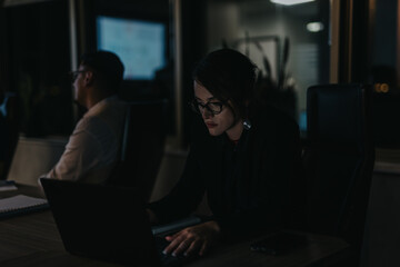 Two business people focused on their work in a dimly lit office during nighttime hours. The setting...