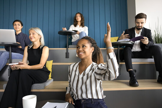 Smiling African female, business training participant raising hand, having creative idea to share, ask to speaker question during meeting or educational seminar in auditorium with diverse teammates