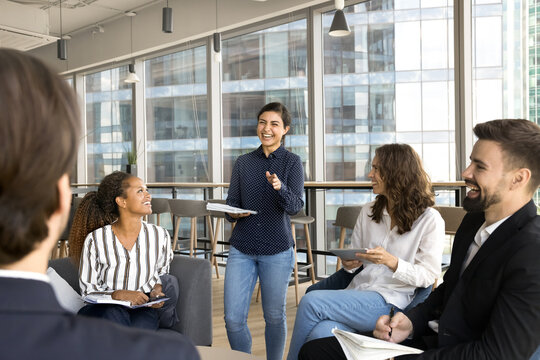 Group of laughing teammates take part in briefing led by young Indian woman, stand in front of colleagues joking, make speech looks hilarious, enjoy communication, friendly relationships at workplace