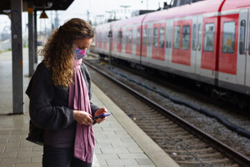 Woman with face mask waiting at platform looking at cellphone in Munich, Germany. Middle aged lady at train station, transportation timetable app, smart phone technology concepts