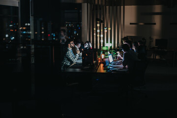 A group of coworkers collaborates on laptops in a dimly lit workspace, focusing on projects. The...