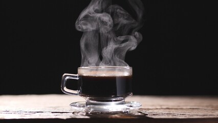 Close up hot coffee clear glass cup, mug and saucer with beautiful steam smoke spins, rising from the cup on old wood table, dark or black background. Concept of coffee and tea, food and drink.