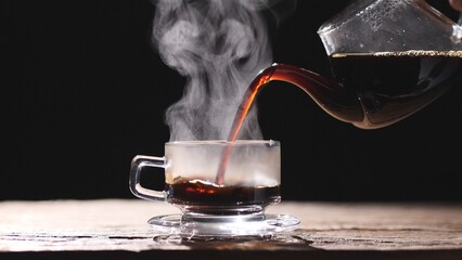 Pouring hot coffee into a clear glass cup, mug and saucer with beautiful steam smoke spins, rising from the cup on old wood table, dark or black background. Concept of coffee and tea, food and drink.