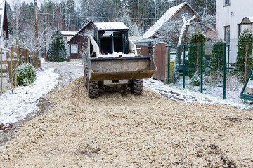 Obraz premium A small construction vehicle spreads gravel on a snow dusted road in a residential area with sloped roof houses, fences, sparse trees, and a wooded backdrop.