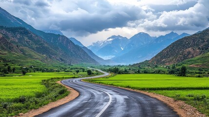 Fototapeta premium Countryside roads nature concept. Serene road winding through lush green fields and majestic mountains under a cloudy sky.