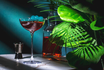 Rob Roy alcoholic cocktail drink with scotch whiskey, sweet vermouth, bitters and cherry. Marble table, blue background with tropical leaves